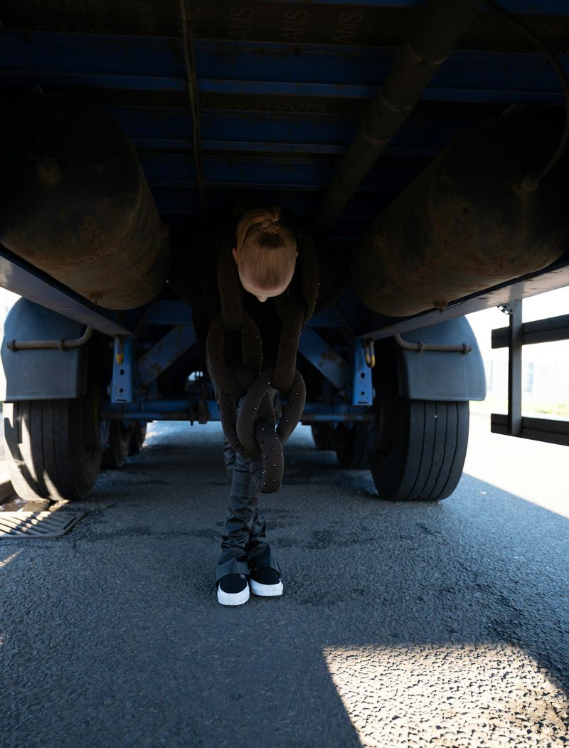 An adult stands underneath the chassis of a semi-truck holding a heavy chain, with the vehicle's tires and undercarriage visible.