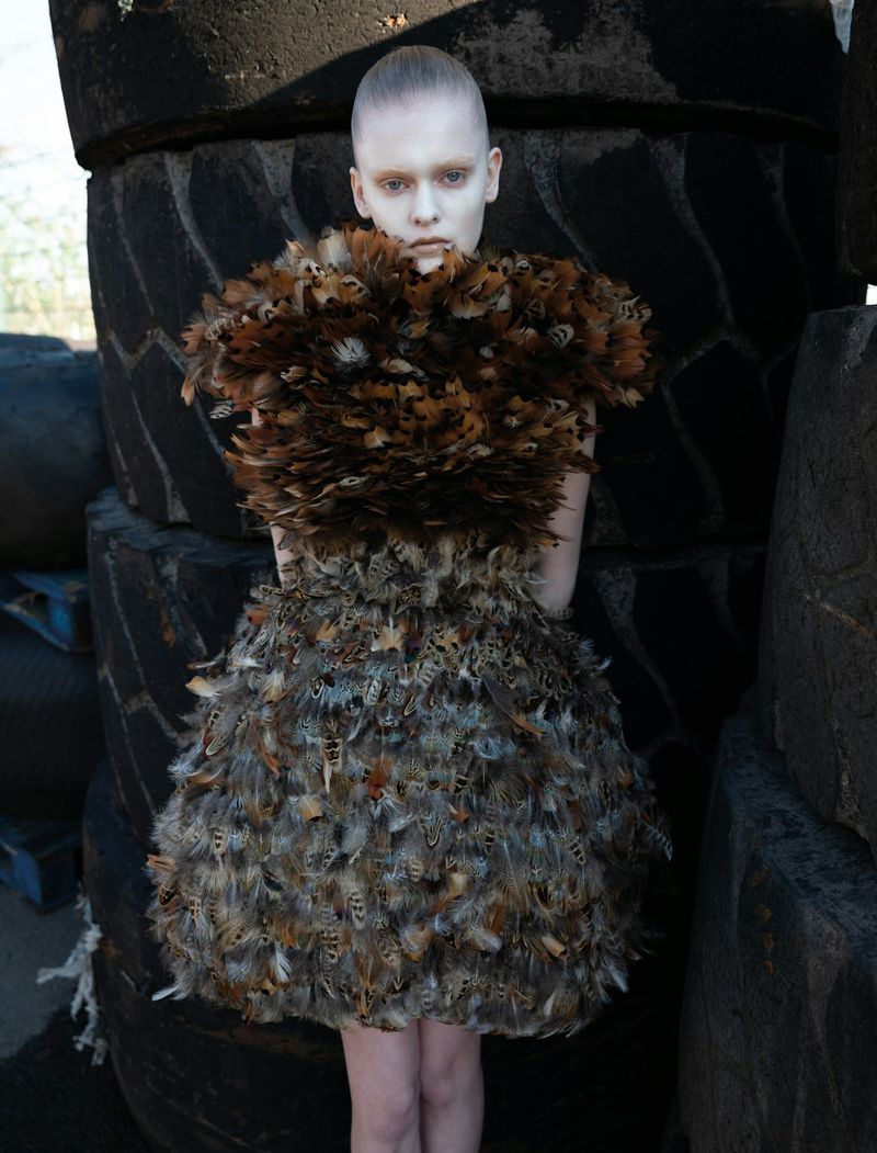 A person wearing a feather dress stands outdoors in front of a stack of tires, creating a high-fashion editorial shot with unusual materials.