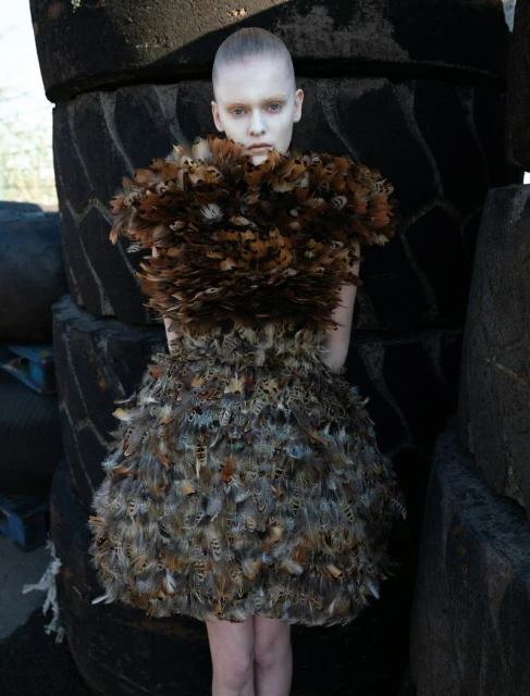 A person wearing a feather dress stands outdoors in front of a stack of tires, creating a high-fashion editorial shot with unusual materials.