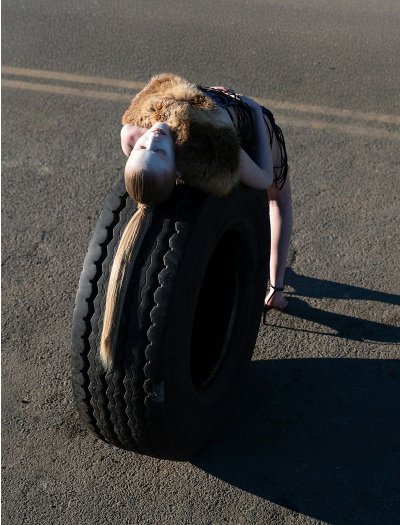 A person laying on a car tire, with their head on top of the tire and their body arched backwards. Pavement and a road are visible.