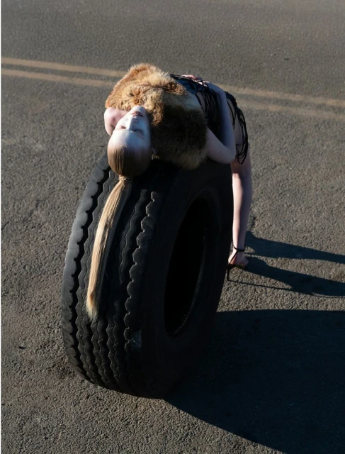 A person laying on a car tire, with their head on top of the tire and their body arched backwards. Pavement and a road are visible.