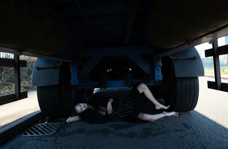 Adult female laying under a semi truck on the asphalt road in black dress and sandals in an outdoor urban daytime environment.