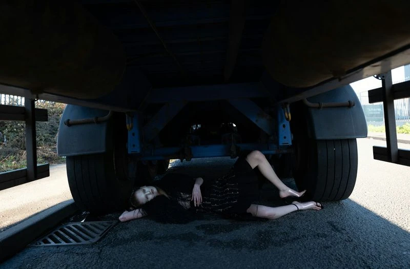 Adult female laying under a semi truck on the asphalt road in black dress and sandals in an outdoor urban daytime environment.
