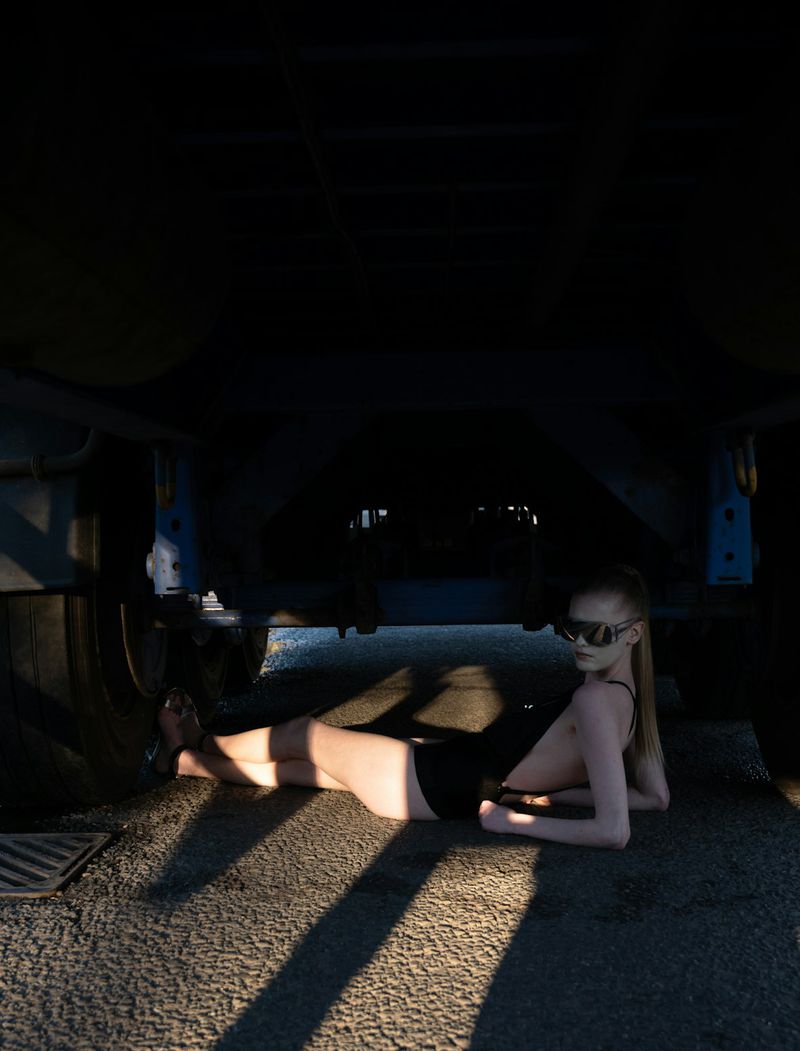 Model wearing dark sunglasses and a black swimsuit lying beneath the undercarriage of a large vehicle outdoors.