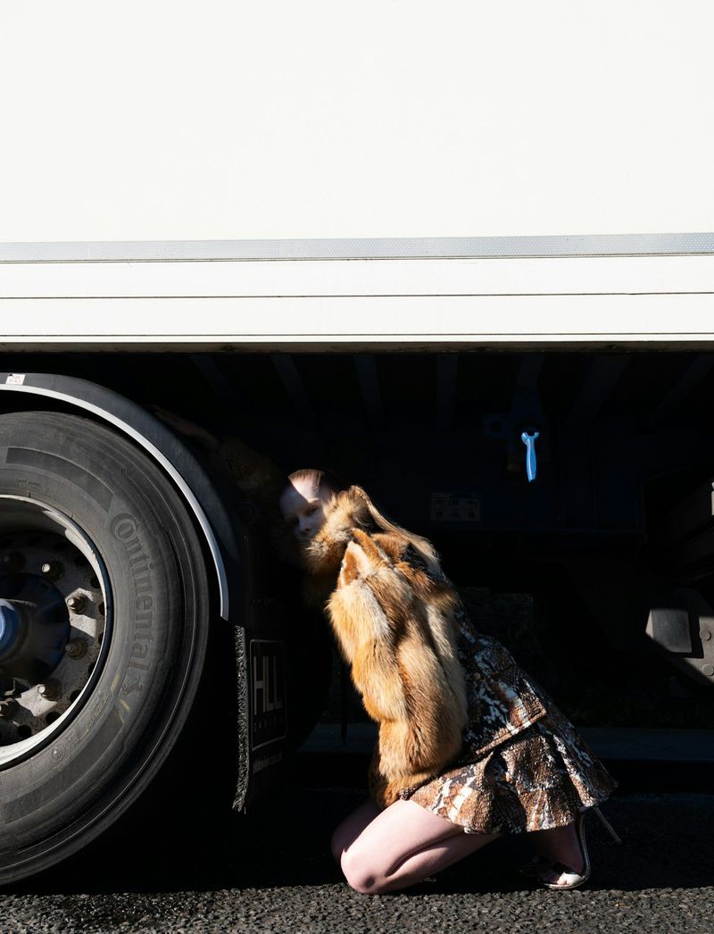 Blonde female in a fur coat and animal print skirt poses kneeling next to the tire of a large white truck in an outdoor setting.