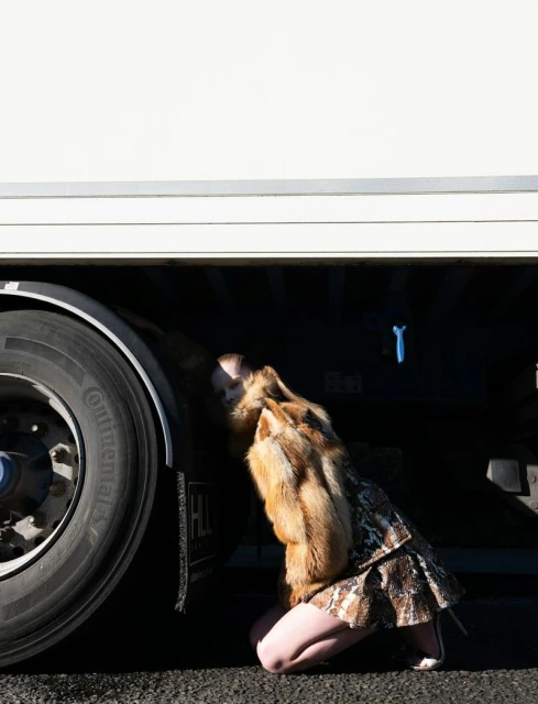 Blonde female in a fur coat and animal print skirt poses kneeling next to the tire of a large white truck in an outdoor setting.