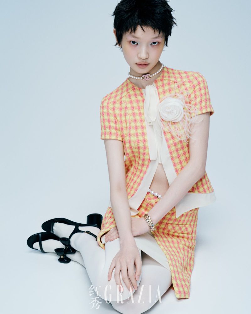Young woman in yellow plaid skirt and jacket, white stockings and black shoes poses in a studio with a light blue background.