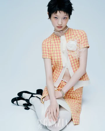 Young woman in yellow plaid skirt and jacket, white stockings and black shoes poses in a studio with a light blue background.