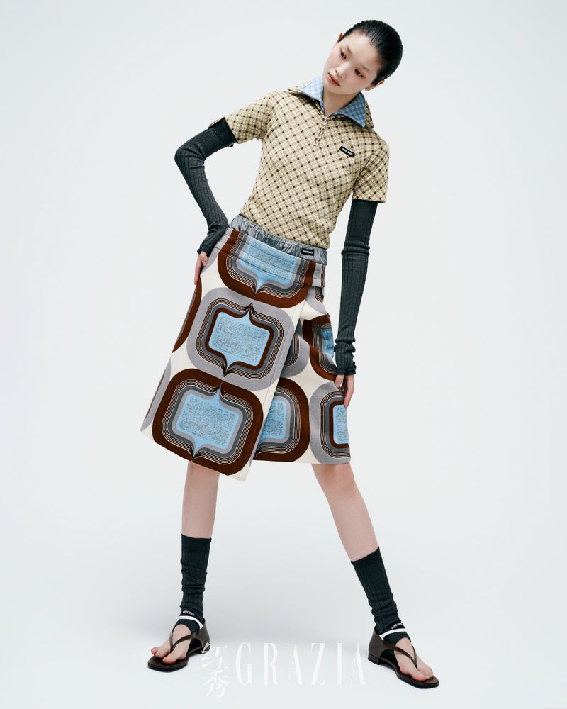 A young adult woman poses in a patterned polo shirt and wrap skirt, paired with arm warmers and sock-sandals on a white background.