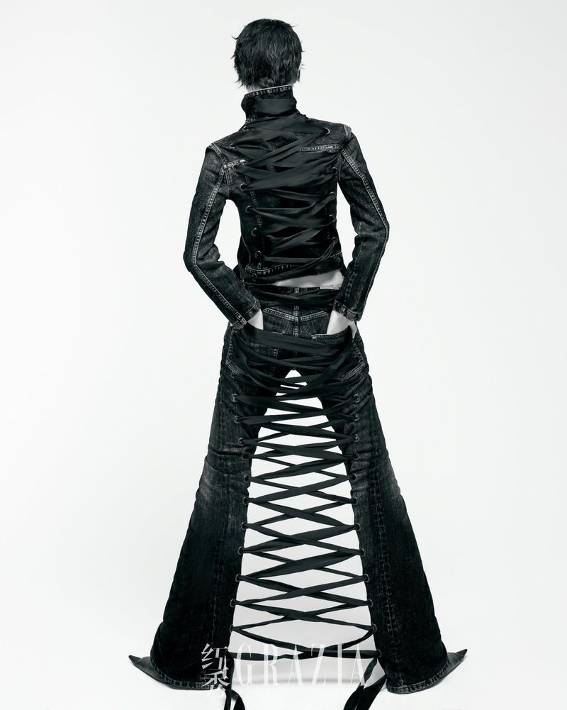 Back view of a person wearing a black jacket and distressed jeans with ribbon details, standing against a stark white background.