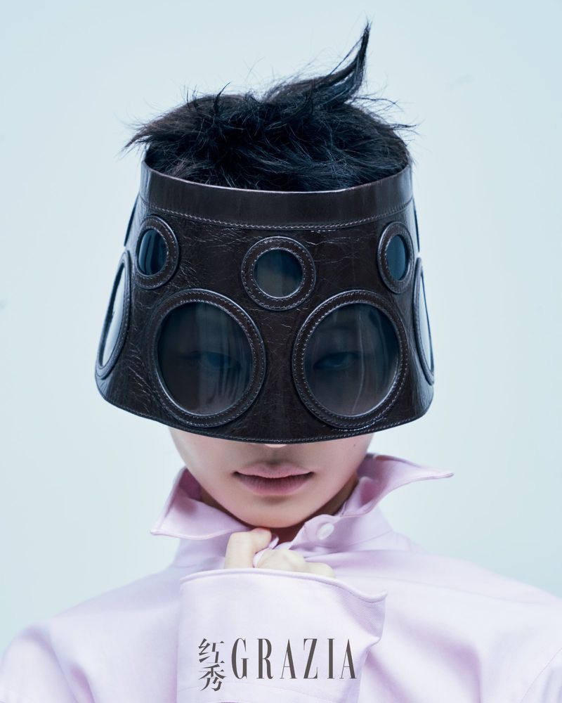 Fashion portrait featuring a person with a dark leather visor with circular cutouts, pale pink shirt, and a Grazia magazine.