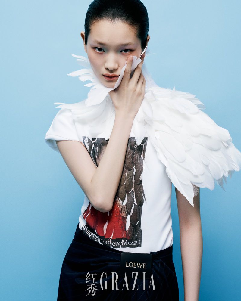 Asian woman poses in a white graphic tee with feathered accents against a bright blue backdrop, fashion forward, high-key lighting.