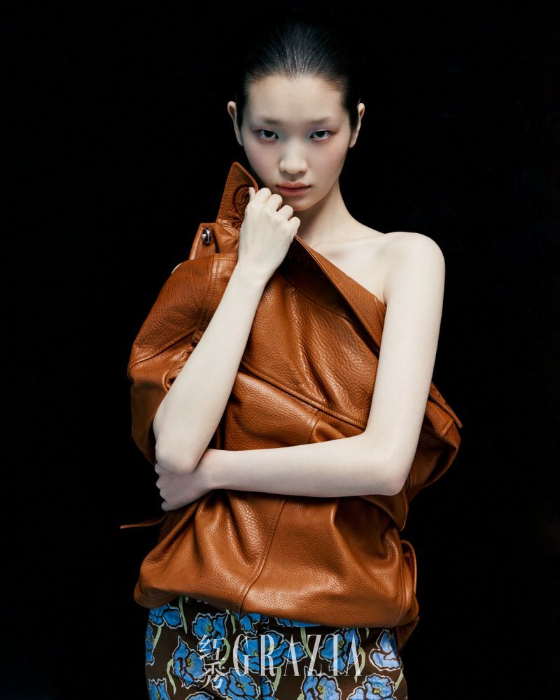 Fashion portrait of an Asian woman with bare shoulders and slicked-back hair, holding a brown leather jacket in front of a black backdrop.