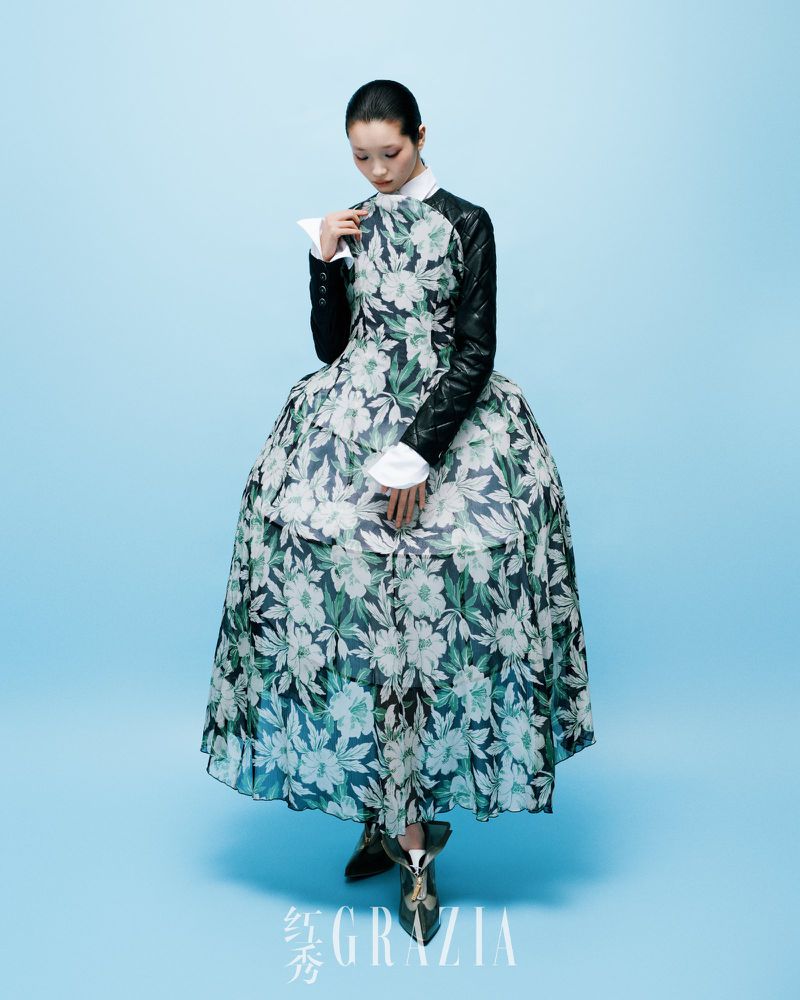 Fashion model poses in a floral gown with black leather sleeves against a light blue backdrop in a studio, showcasing contemporary fashion.