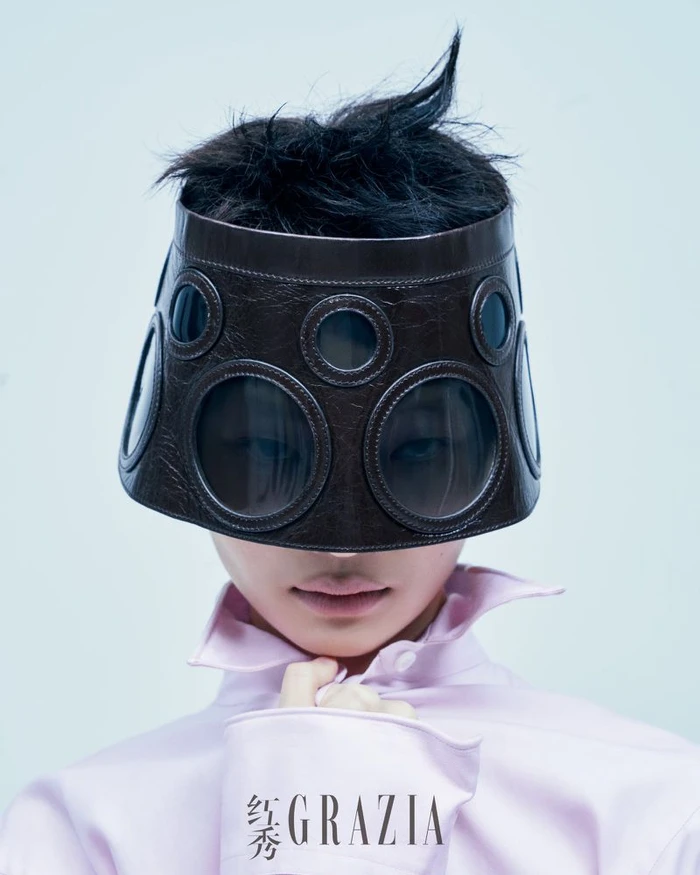 Model wearing a dark brown sculptural visor hat with circular portholes and a light pink shirt against a pale background