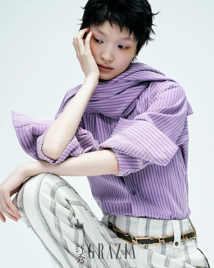 Model with dark short hair wearing a purple striped shirt and white patterned pants against a white background