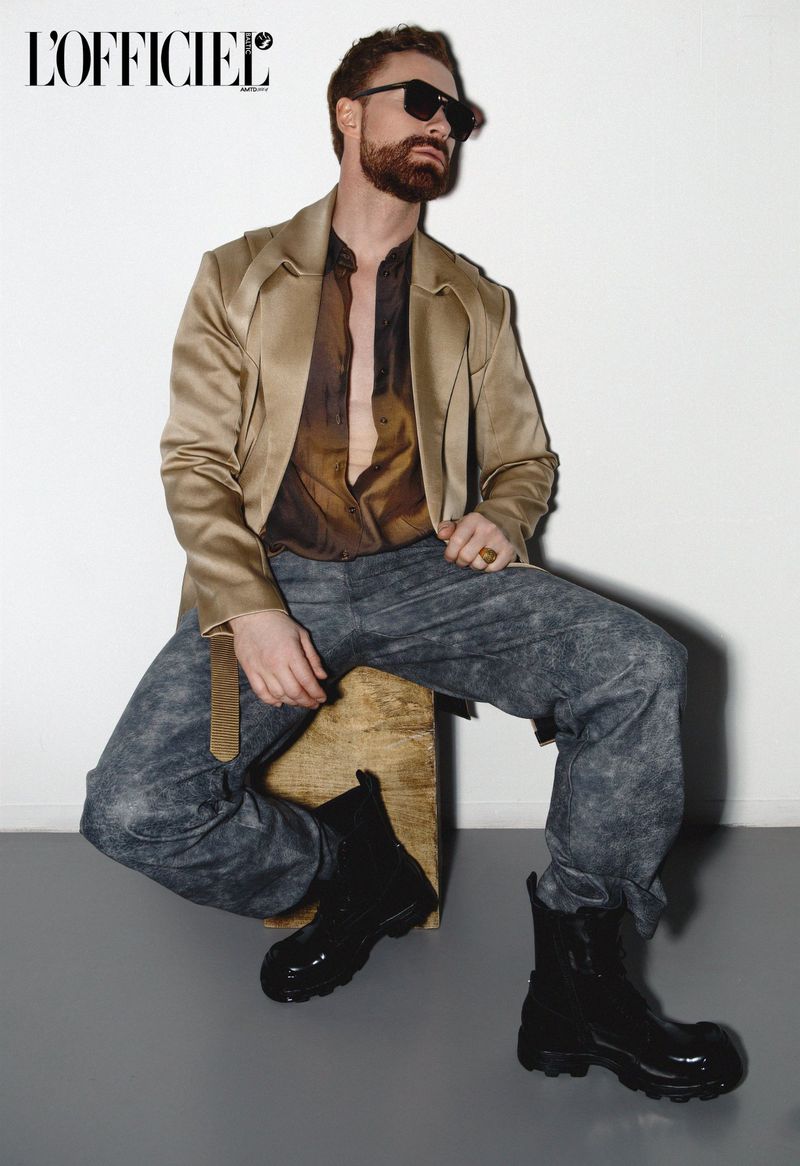 A bearded, adult man in a jacket and sunglasses sits on a wooden crate wearing black boots in a fashion shot on a grey and white background.