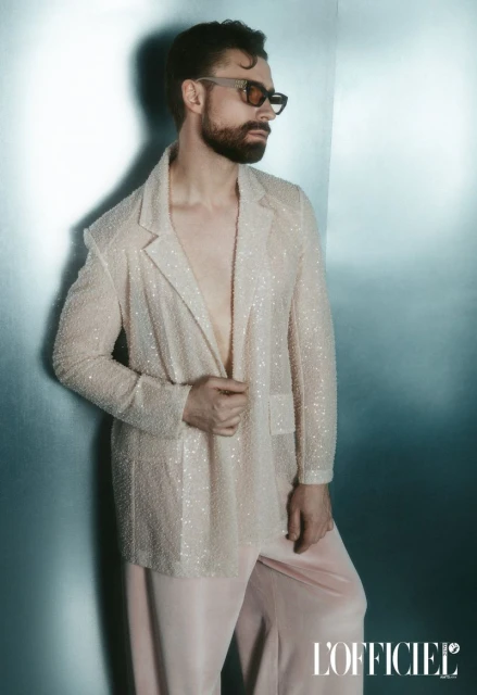 Man with beard and sunglasses poses in shimmering blazer and pink pants against a textured wall, studio lit, fashion portrait.