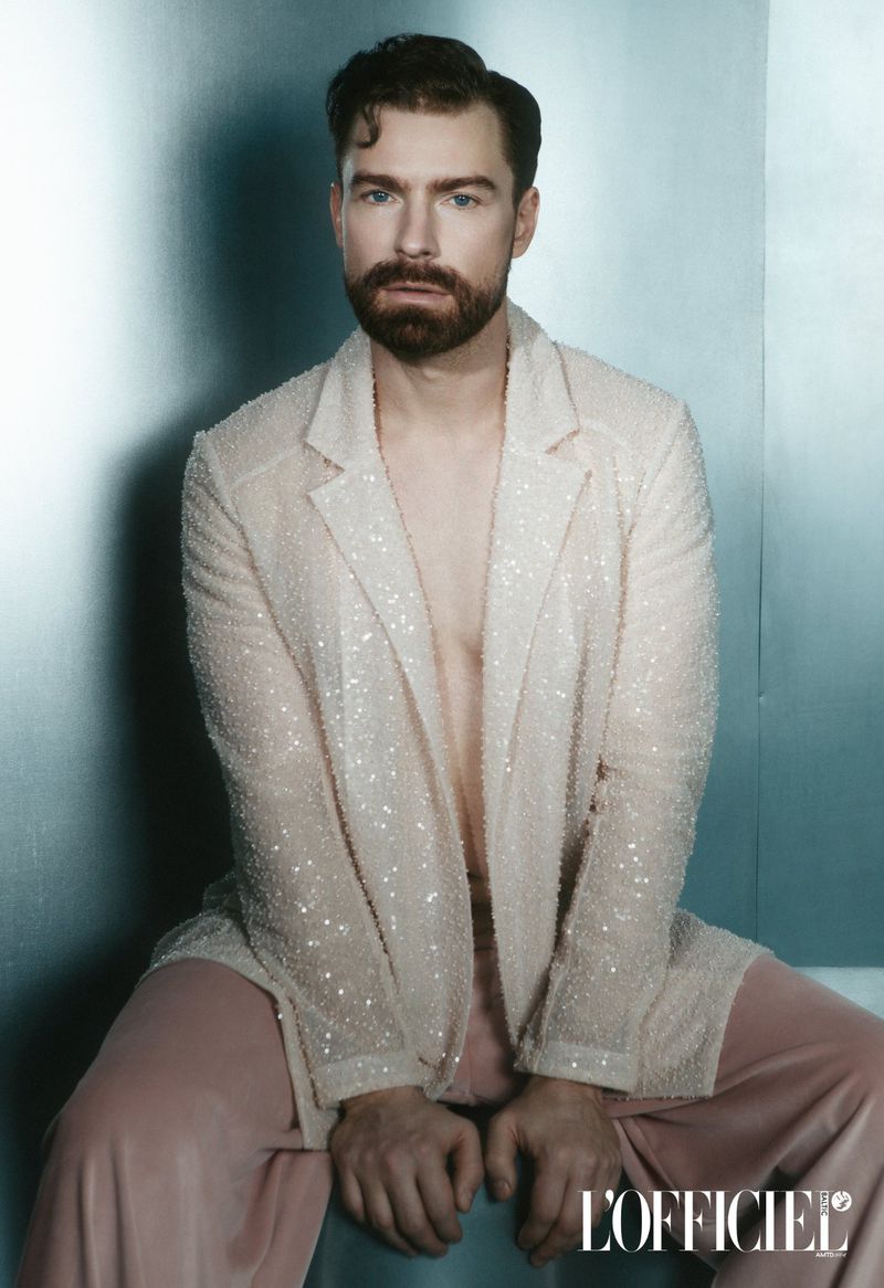 Bearded man poses indoors wearing a light-pink sequined blazer and pink pants in a fashion editorial shot on a blue-gray background.