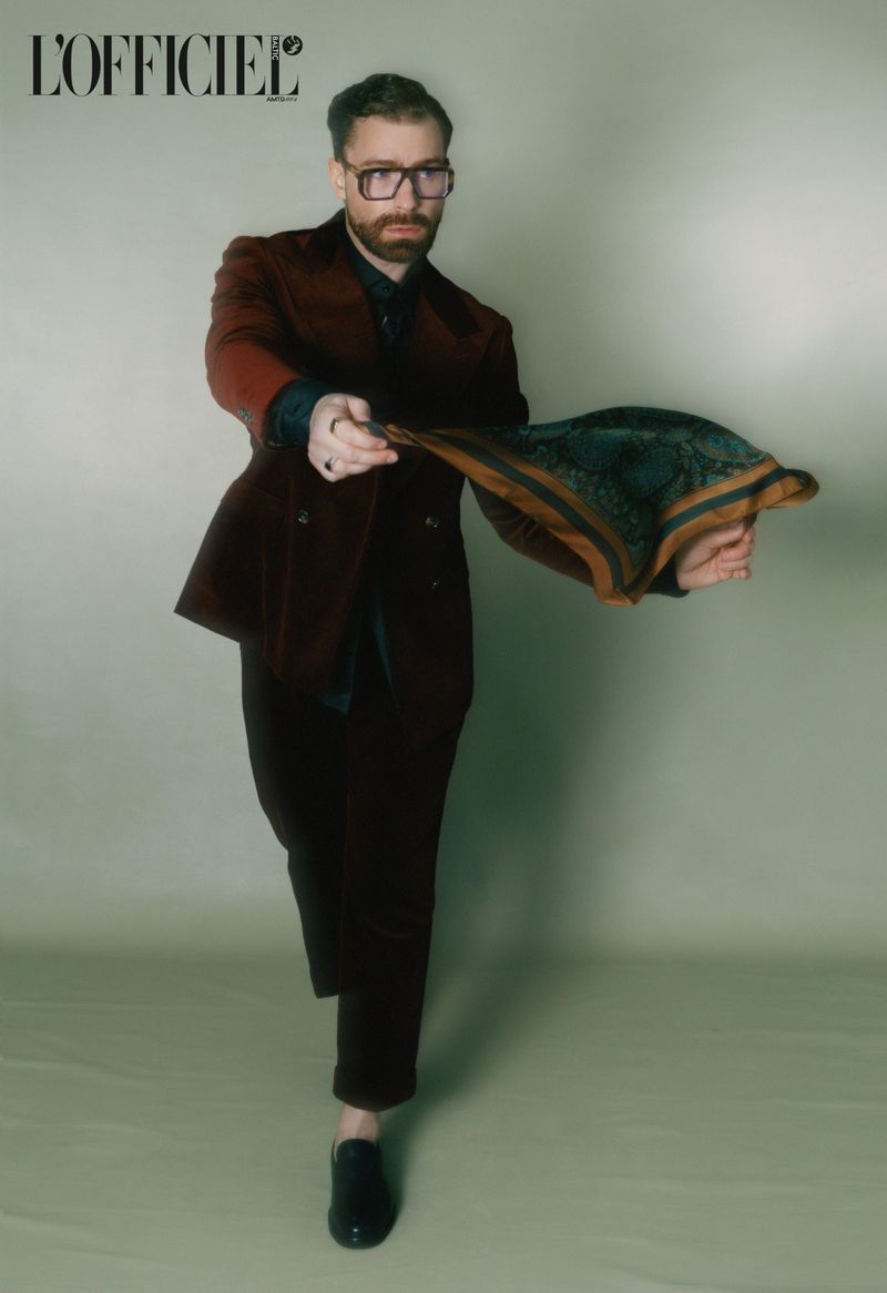 Man in a maroon suit holding a silk scarf, glasses, indoors, fashion editorial portrait for L'Officiel magazine, studio lighting, neutral background.