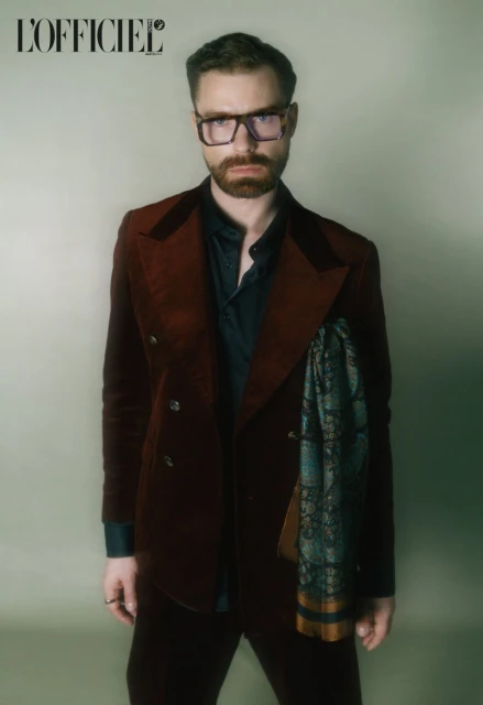 Portrait of a man with a beard wearing glasses, a burgundy suit, and a patterned scarf in a studio setting with diffused lighting.