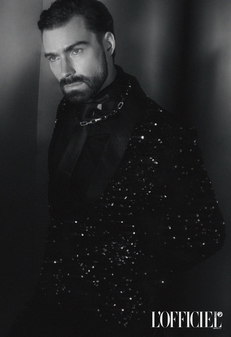 Black and white portrait of a bearded adult male wearing a sequined jacket in a low-key lighting environment against a dark wall.
