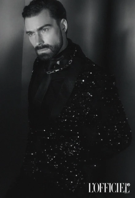 Black and white portrait of a bearded adult male wearing a sequined jacket in a low-key lighting environment against a dark wall.