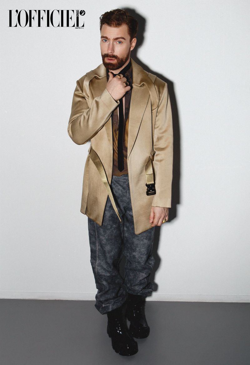 Portrait of a red-haired man in a tan coat and grey patterned pants standing in front of a white wall, shot at eye-level in a studio.