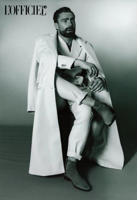 A stylish man with a beard, wearing a white coat, jacket, pants, and boots, poses in a studio setting with the 'L'Officiel' logo visible.