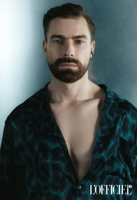 Portrait of a bearded adult male in a patterned shirt against a mottled blue-gray backdrop, shot in a photo studio with L'Officiel logo.