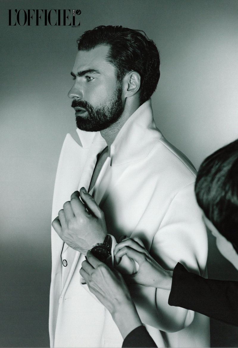 Profile shot of a bearded man in a white jacket, being adjusted by an unseen assistant against a light gray background.
