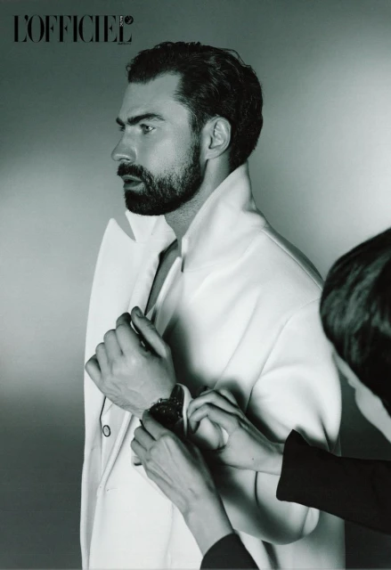 Profile shot of a bearded man in a white jacket, being adjusted by an unseen assistant against a light gray background.