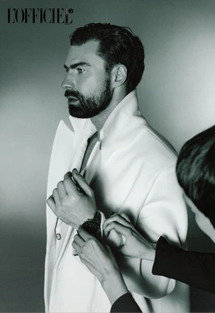 Profile shot of a bearded man in a white jacket, being adjusted by an unseen assistant against a light gray background.