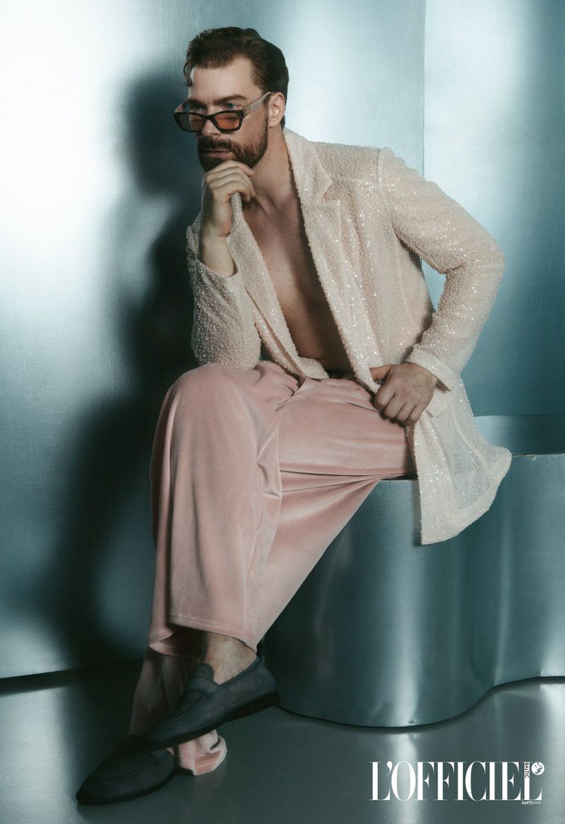 Man with beard wearing sunglasses, textured cream jacket, and pink trousers, seated against a metallic background