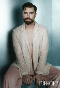 Man with beard wearing a glittering cream blazer and pink trousers against a blue metallic background