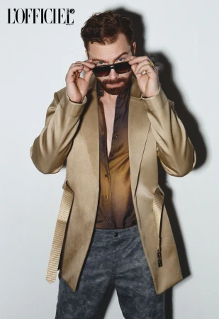 Man adjusting sunglasses wearing a gold blazer and patterned trousers against a plain white wall