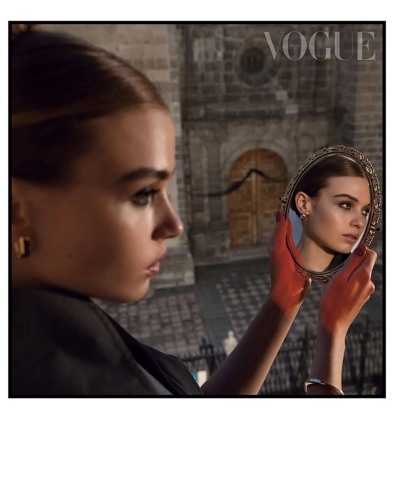 Young woman in profile holding vintage mirror reflecting her face against a background of stone architecture and a 'Vogue' masthead.