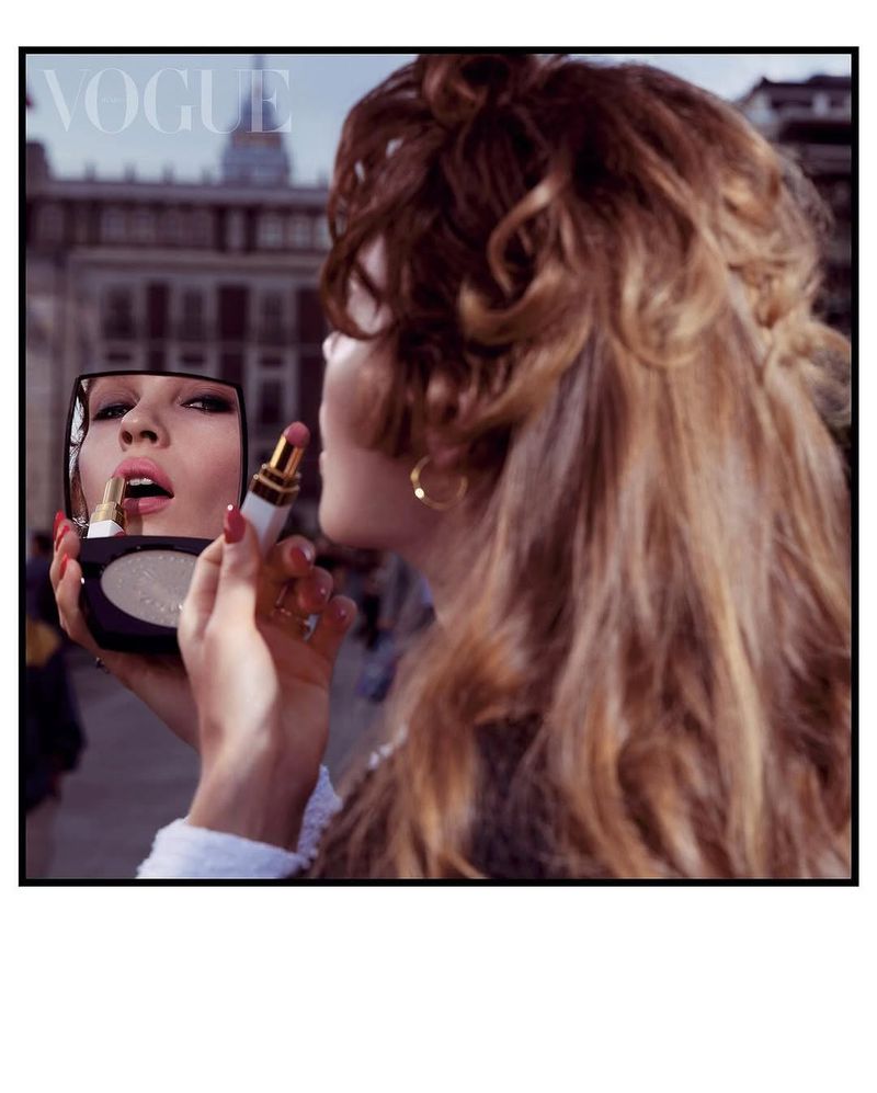Woman applies lipstick with compact mirror outdoors in city; Vogue magazine masthead visible in the upper left of the image.