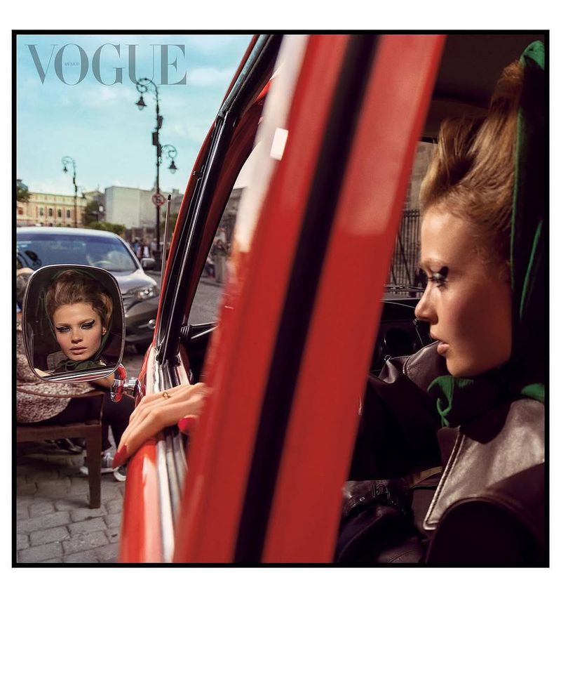Vintage-style portrait of an adult female driving a red car in a European city, reflected in the side mirror, with Vogue logo.