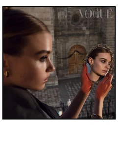 Model holding an ornate oval hand mirror reflecting her face, set against a stone architectural building exterior