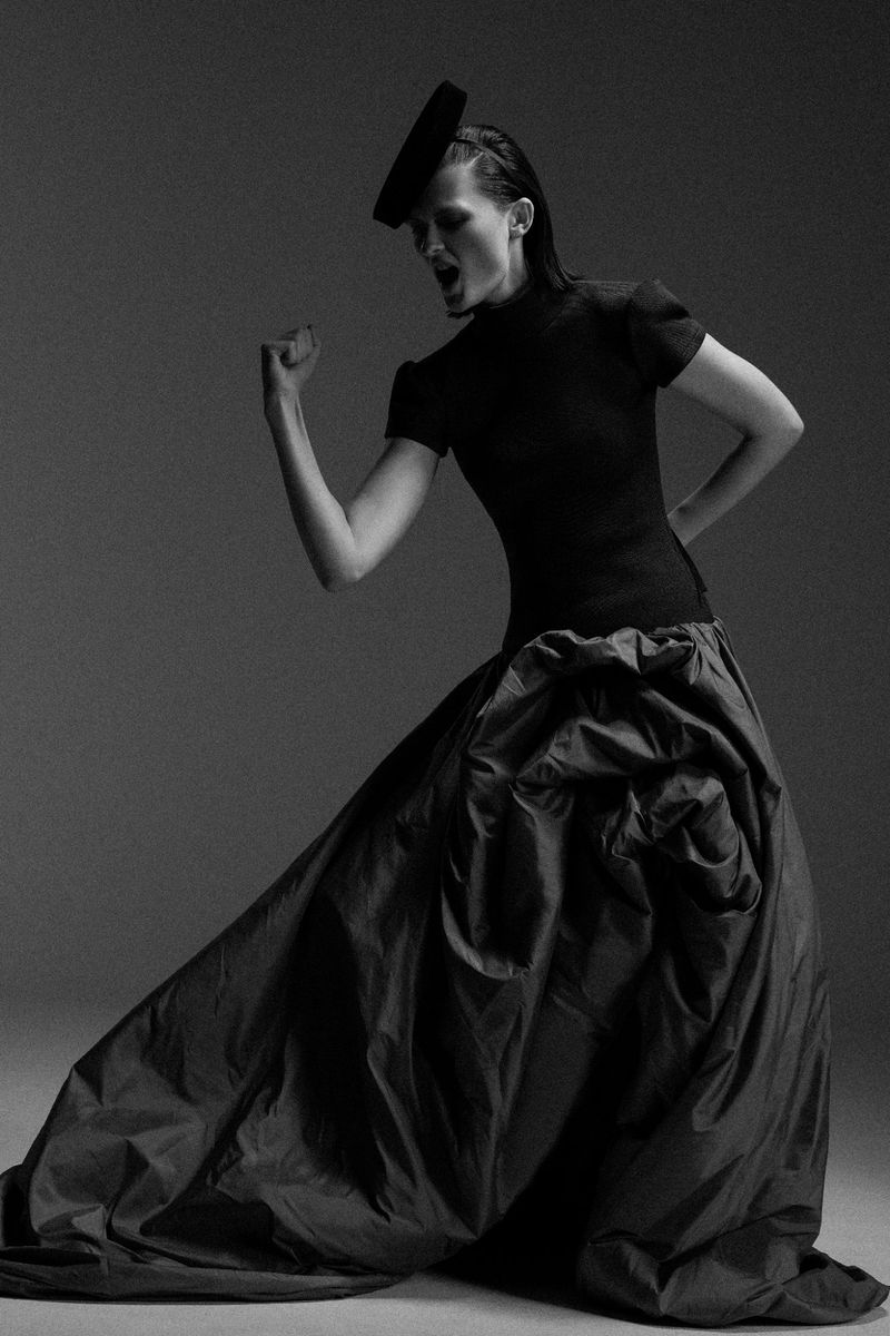 Dramatic black and white fashion shot: Woman in oversized taffeta skirt and fitted top with theatrical pose against a grey backdrop.