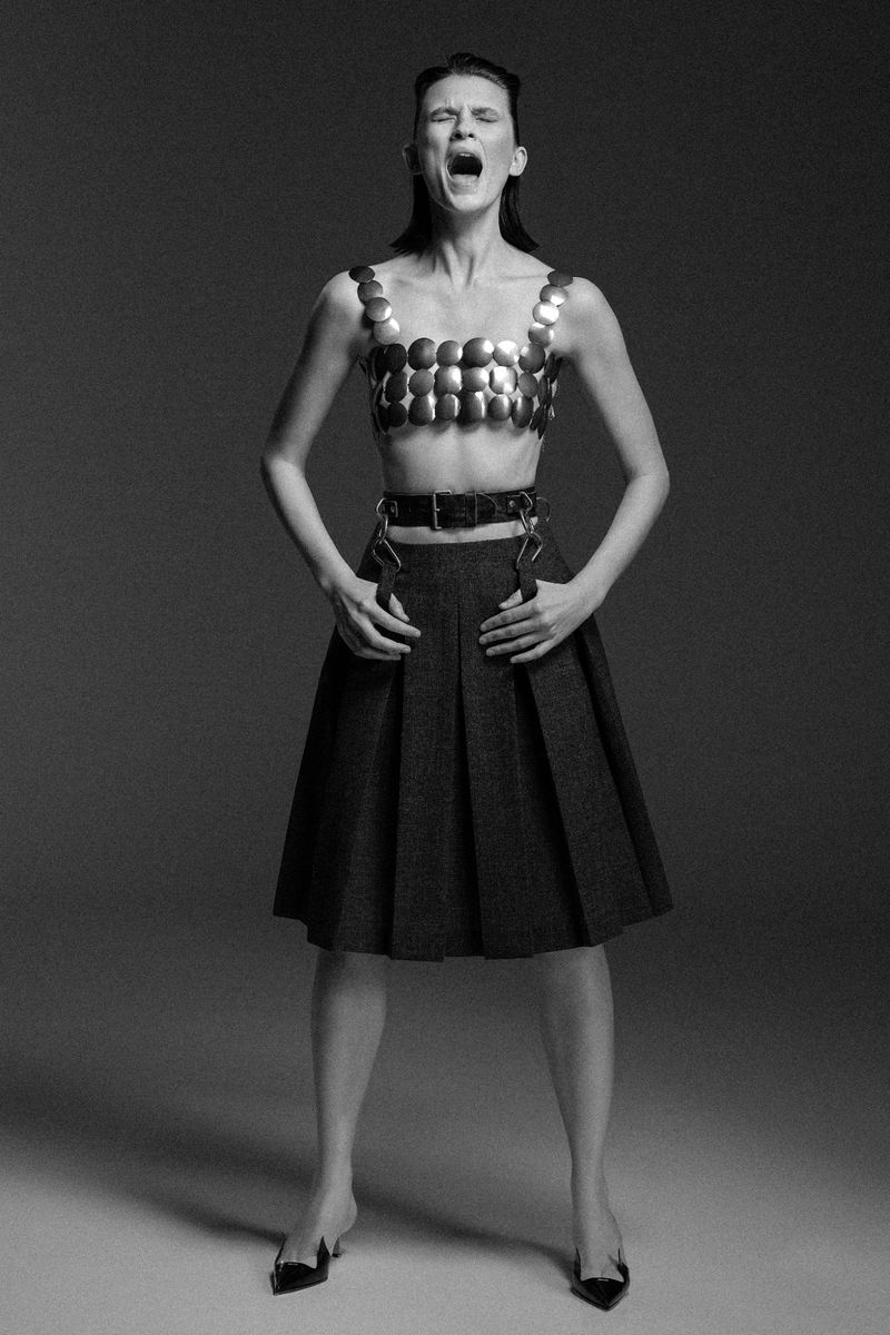 Monochrome shot of a woman in a metal top and skirt, dramatically posing with an open mouth in a studio against a grey backdrop.