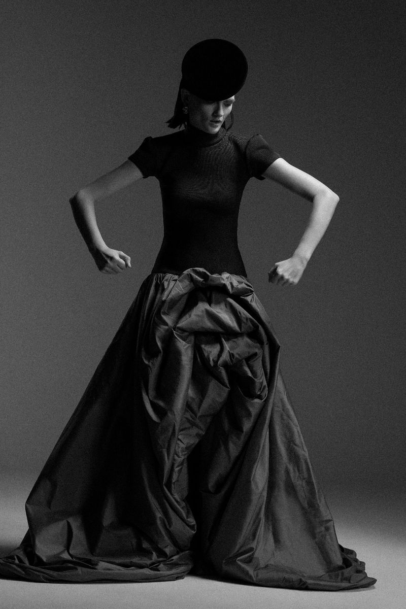 Black-and-white fashion shot: a woman in a sculptural dress with a wide skirt and fitted top poses with arms bent in a photo studio.