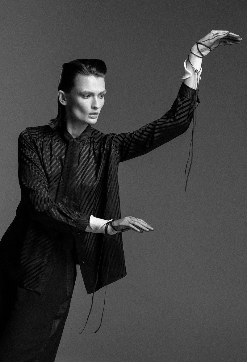 Monochrome fashion shot featuring a model in a striped shirt and dark skirt, posing with arms raised in a minimalist studio setting.
