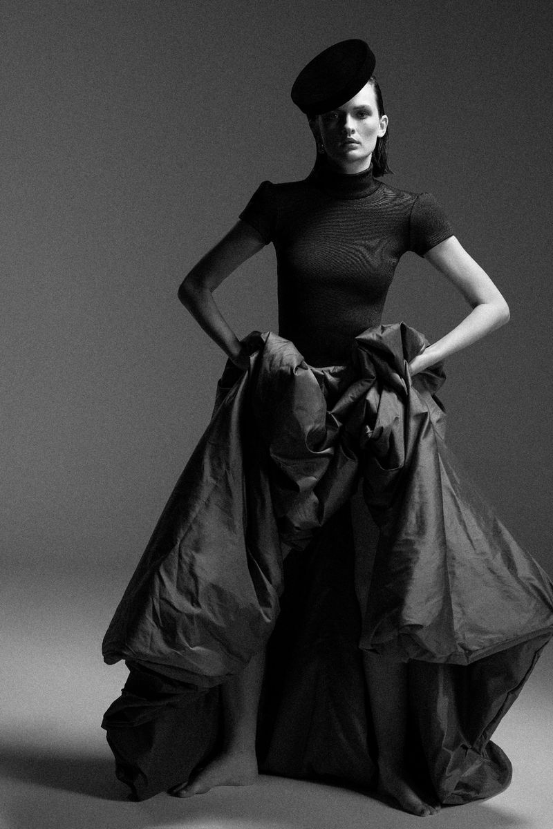 A slender model poses in a black, short-sleeved top, large black skirt and matching hat against a neutral studio backdrop.