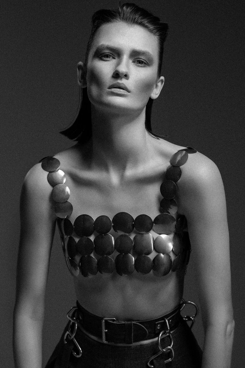 Monochrome portrait of a fair-skinned adult woman with dark hair wearing a metallic disc top and dark belt. Studio lighting, gray backdrop.