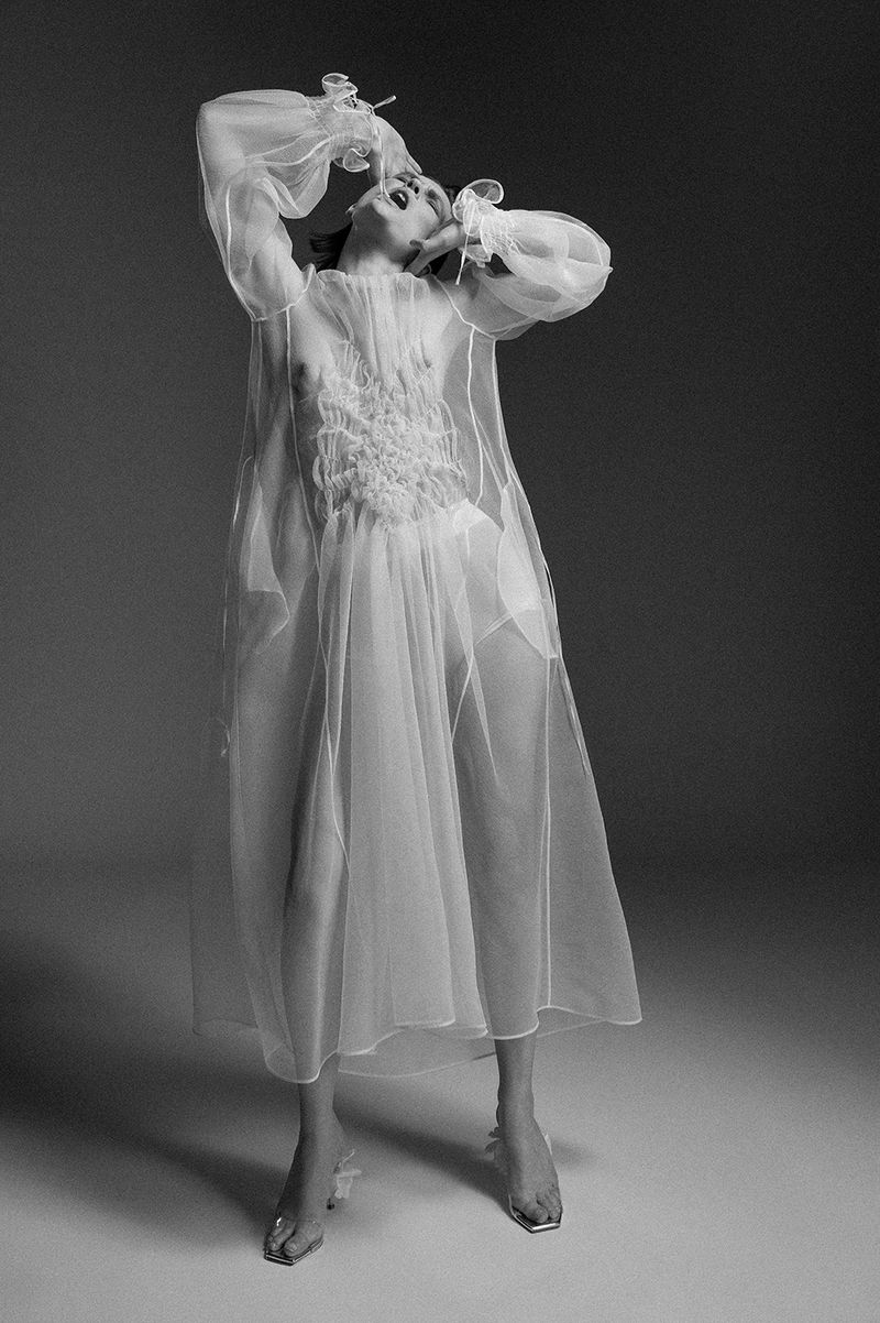Black and white studio shot of a person wearing a translucent dress, looking upwards with hands near face, against a neutral backdrop.