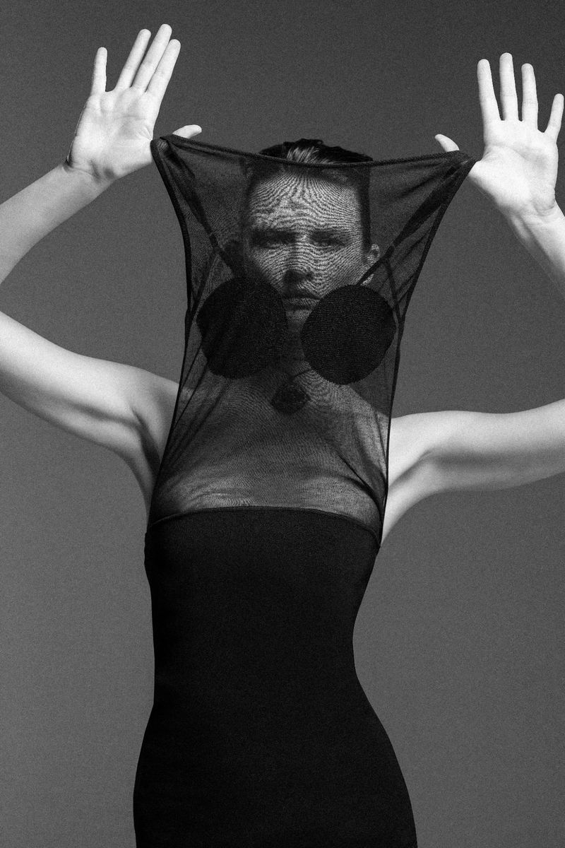 Monochrome fashion portrait of a person in a black dress holding up a mesh garment over their face with both hands in a studio setting.