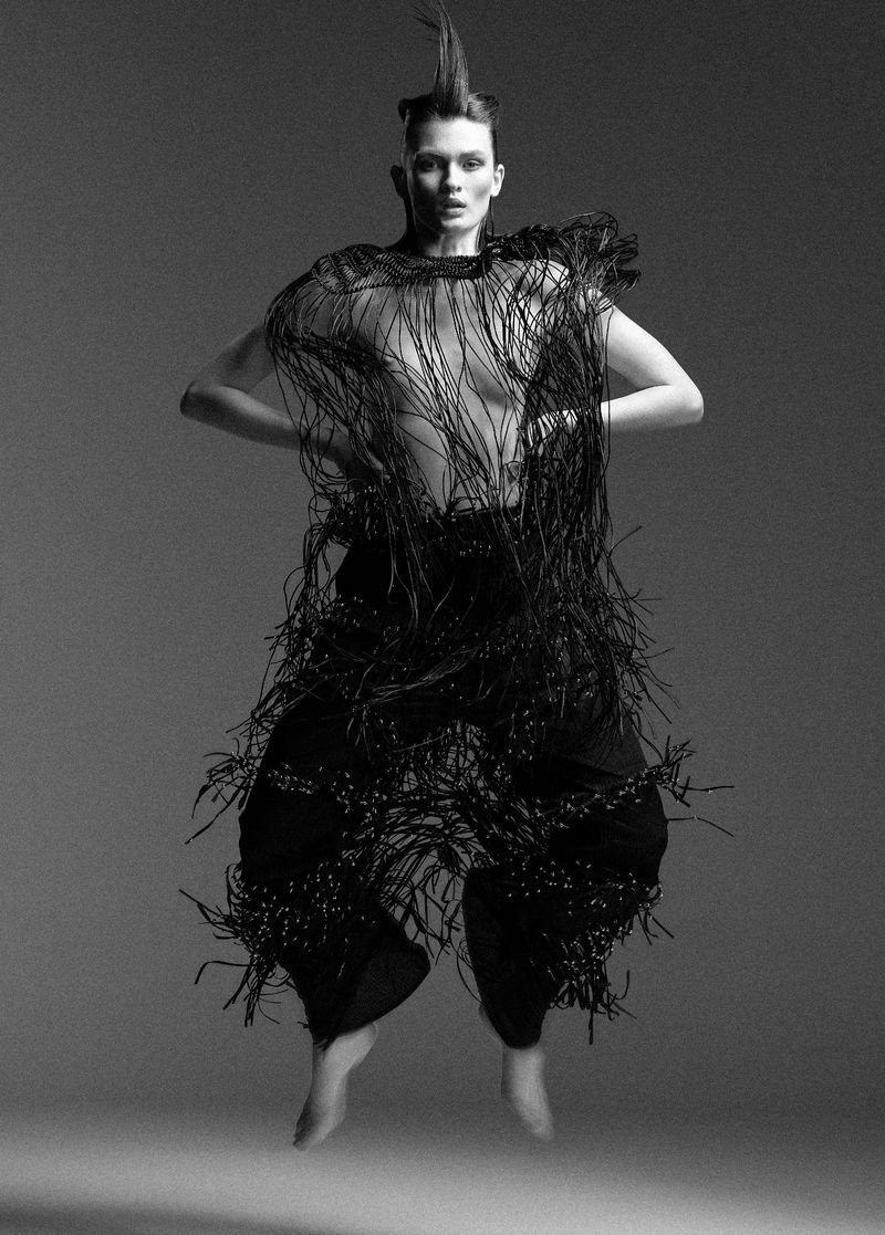 A person jumps in the air wearing a fringed black top and pants in a studio shot, barefoot, with a light grey background.