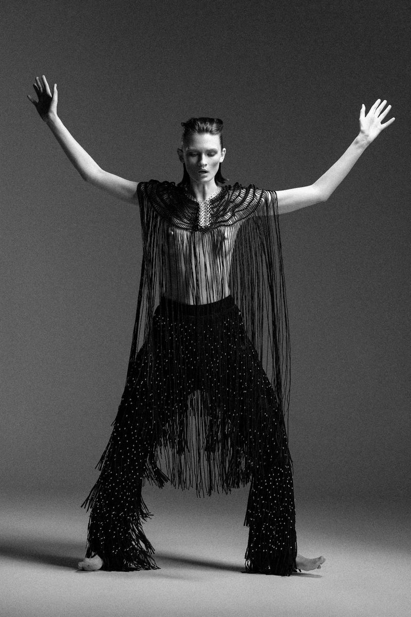 Monochrome studio shot of a female fashion model in fringed cape and trousers, striking a pose with raised arms against grey background.